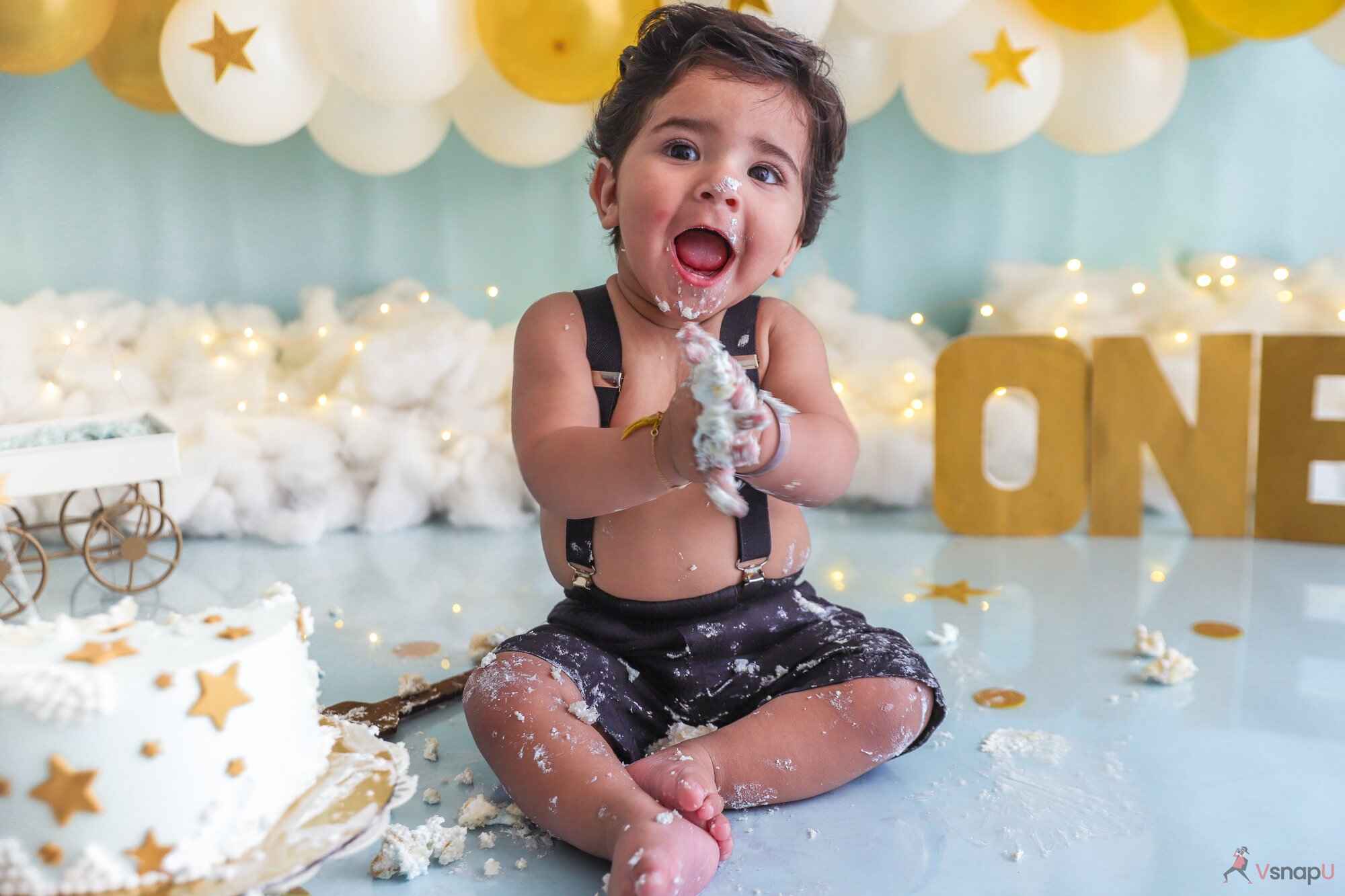 A sweet one-year-old baby laughing with cake on their face, surrounded by golden and white balloons during their baby photoshoot.
